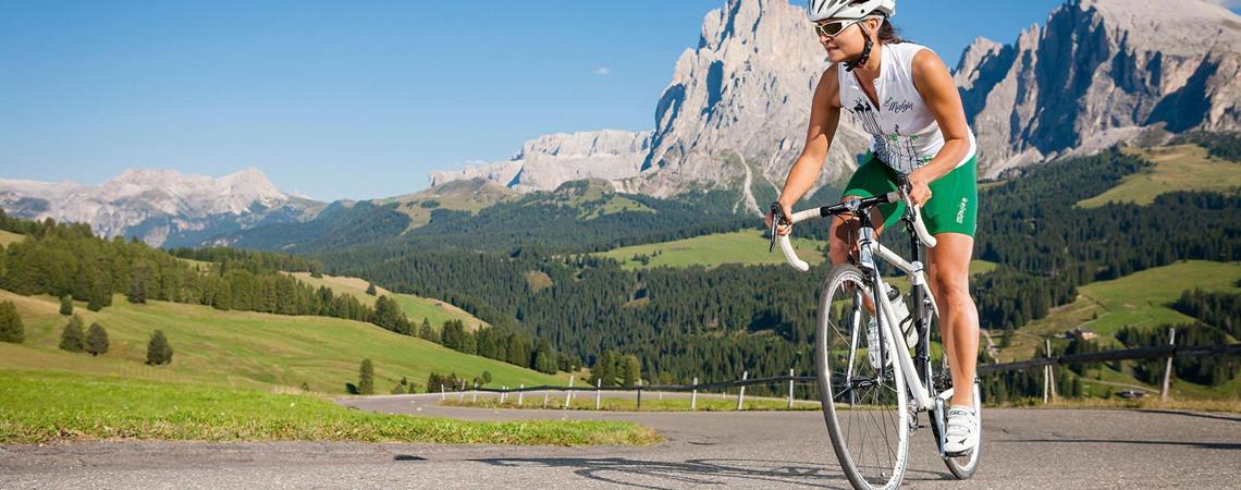 Donna con casco pedala su una strada di montagna sotto il sole, circondata da colline verdi.