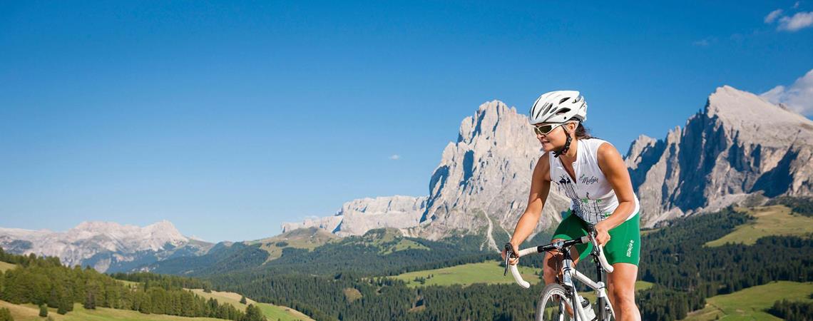 Woman with helmet cycling on a mountain road in sunshine, surrounded by green hills.