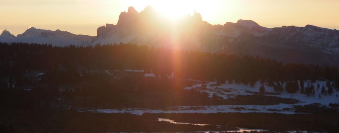 Alba splendente dietro montagne innevate con cielo rosato e dorato e paesaggio invernale coperto di neve in primo piano.