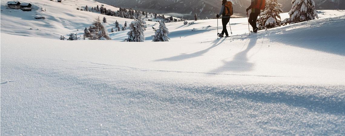 Two snowshoe hikers walk through a snowy mountain landscape at sunset.