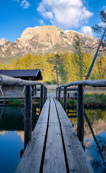 Holzsteg über einen ruhigen See mit Blick auf eine Hütte und den imposanten Schlern