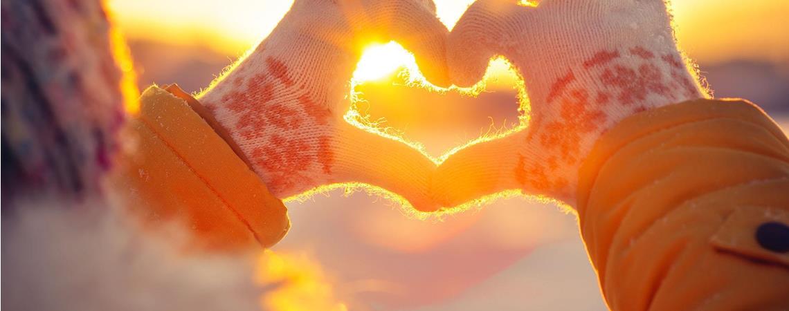 Person mit Winterhandschuhen formt ein Herz mit den Händen vor der untergehenden Sonne.