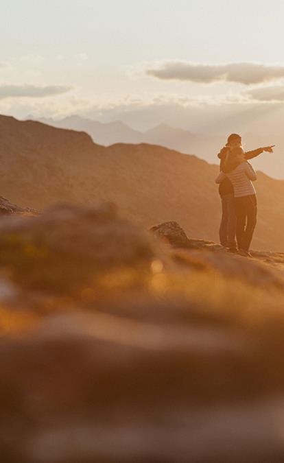 Coppia ammira il panorama alpino al tramonto, una persona indica l'orizzonte.