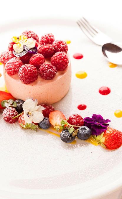 Dessert ai lamponi con frutti di bosco freschi, fiori commestibili e salsa alla frutta su piatto bianco, accompagnato da un bicchiere di vino rosso.