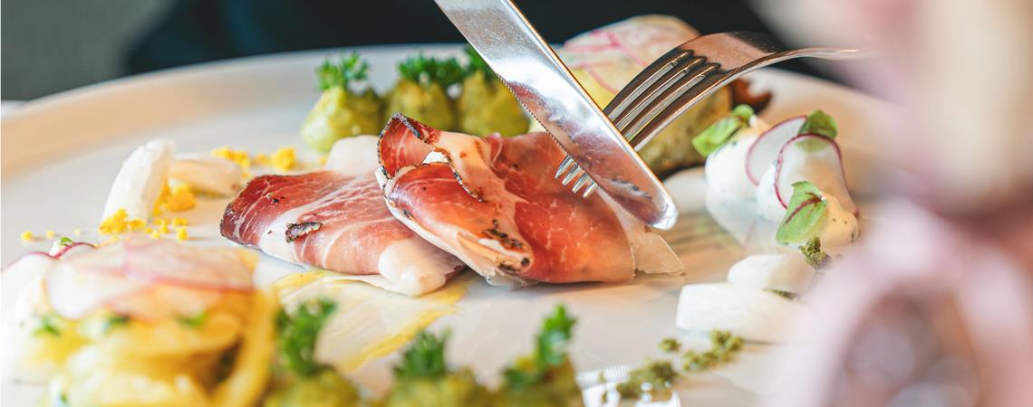 Gourmetgericht mit Südtiroler Speck, Gemüse und Cremes, das mit Messer und Gabel serviert wird.