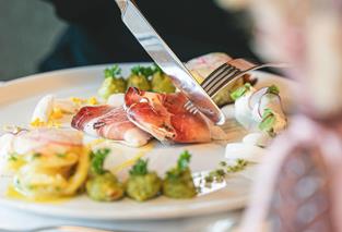 Gourmetgericht mit Südtiroler Speck, Gemüse und Cremes, das mit Messer und Gabel serviert wird.