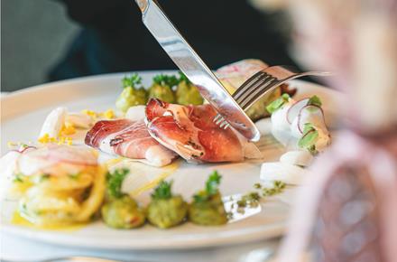 Gourmetgericht mit Südtiroler Speck, Gemüse und Cremes, das mit Messer und Gabel serviert wird.